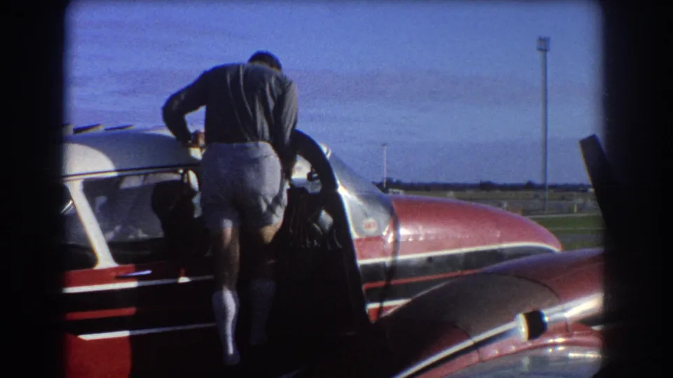 The man begins climbing into the small aircraft, with the open door, wing edge, and runway lighting visible nearby.