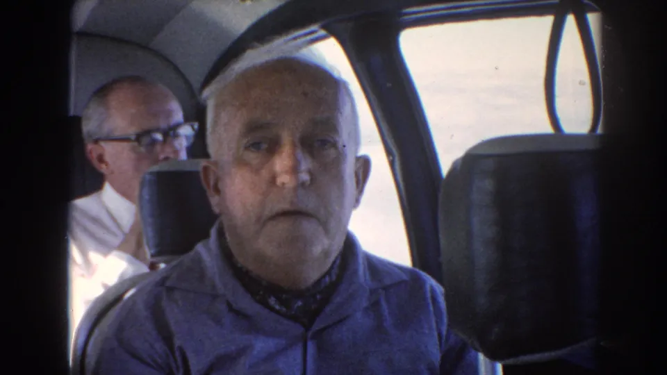 Inside the aircraft cabin, an older man faces forward while another seated man is visible behind him near the window.