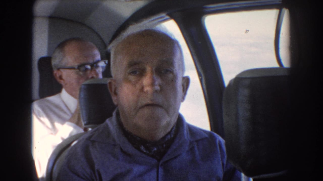 Older man in a blue shirt seated inside a vehicle cabin with another man behind him.