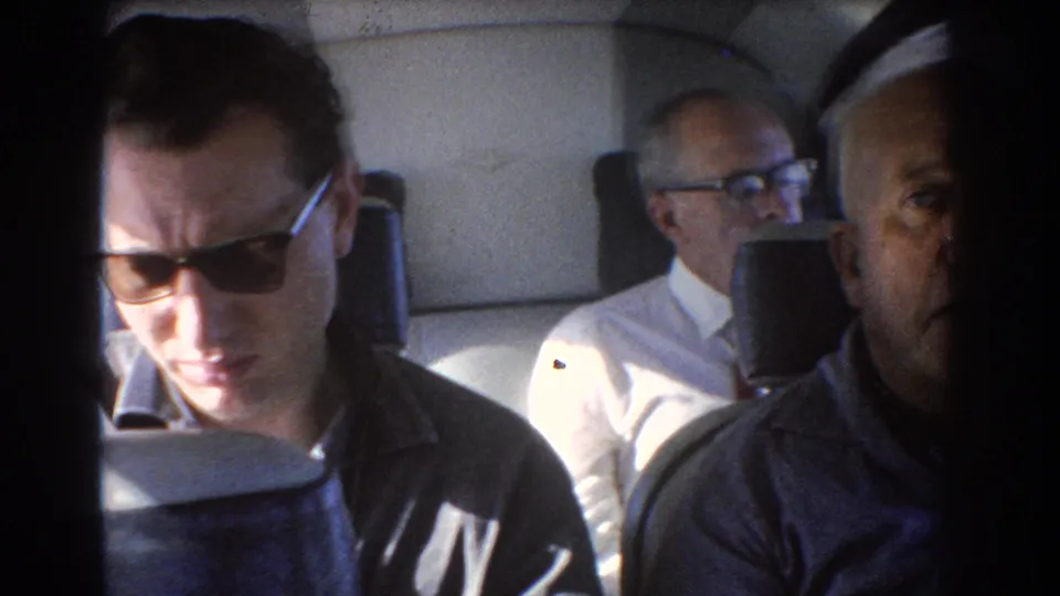 1967 Perth home-movie frame showing a younger man in sunglasses looking down, with older passengers partly visible behind and beside him.