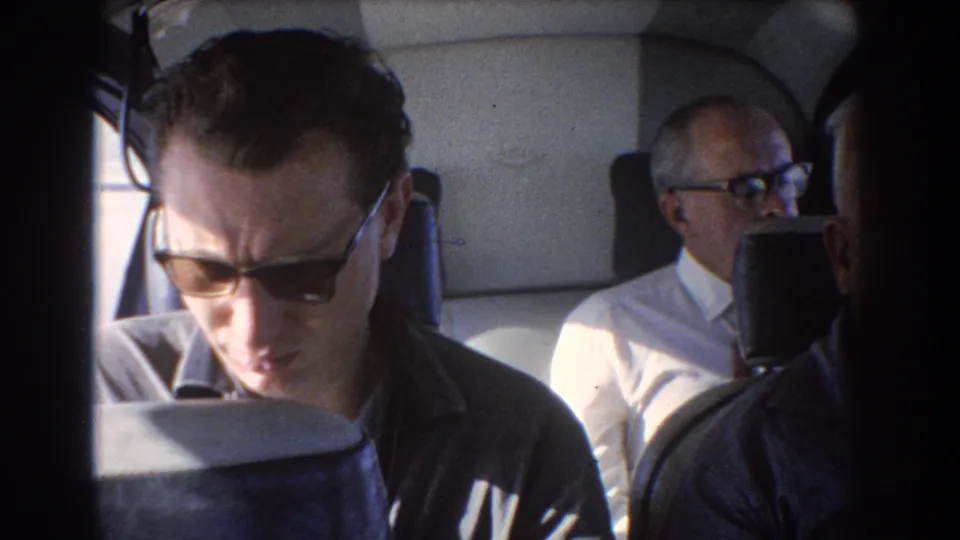 1967 Perth home-movie frame of a younger man in sunglasses seated in the foreground, with an older bespectacled passenger behind him.