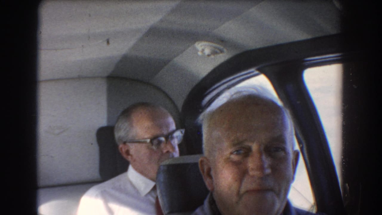 Two older men seated inside an airplane cabin, one facing the camera and one behind him in glasses.