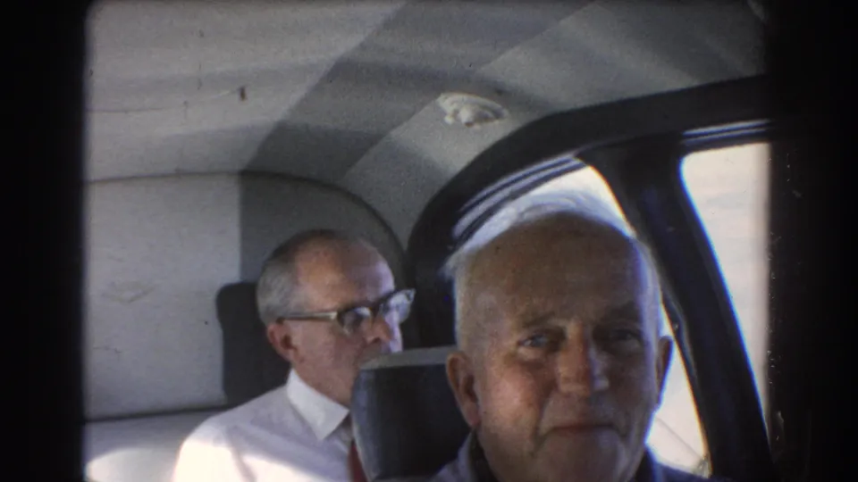 1967 Perth, Australia 8mm frame showing two older men in an airplane cabin, with daylight through the window and dark seatbacks.