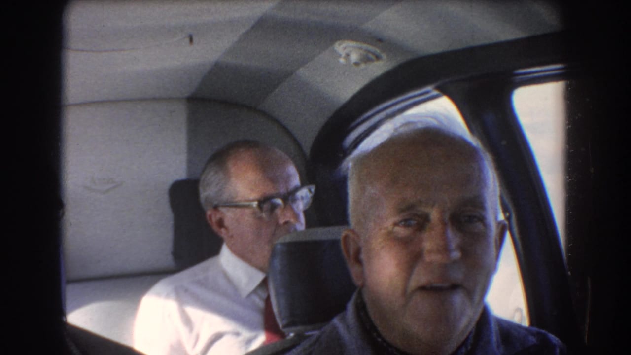 Two older men sit by airplane cabin windows, with the foreground man turned toward the camera.