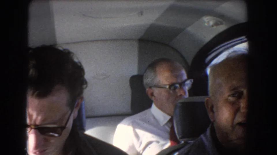 1967 Perth, Australia 8mm frame showing three adult men in an airplane cabin, with a sunglasses-wearing man close to the camera.
