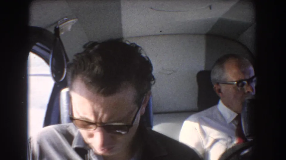 1967 Perth, Australia home-movie frame with a sunglasses-wearing passenger leaning forward and an older man seated behind in the aircraft cabin.