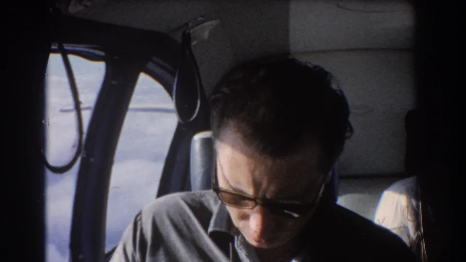 1967 Perth, Australia home-movie frame of a passenger reading beside an aircraft window with cabin structure in view.