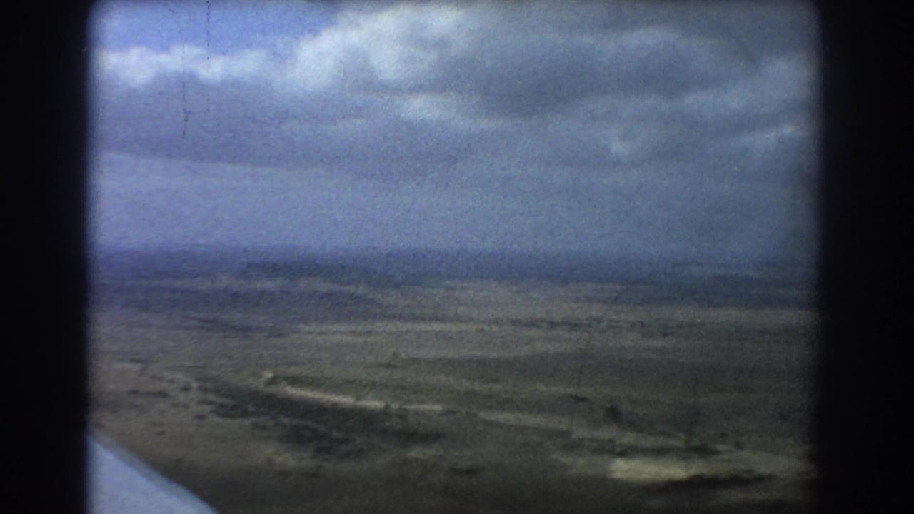 Airplane wing edge above a flat landscape under dark clouds.