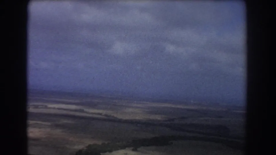 1967 airplane-window footage over flat Perth-area plains, with a low horizon, clouded sky, muted colors, and visible 8mm grain.