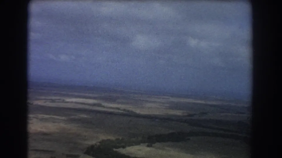 1967 Perth-region home-movie aerial view over arid terrain, with low hills under a cloudy sky and dark film-frame borders.