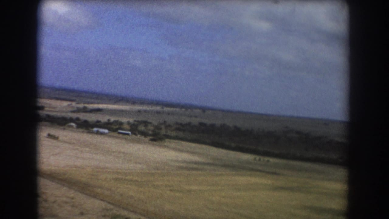 Sandy open field with sparse greenery, distant structures, and blue-gray sky.