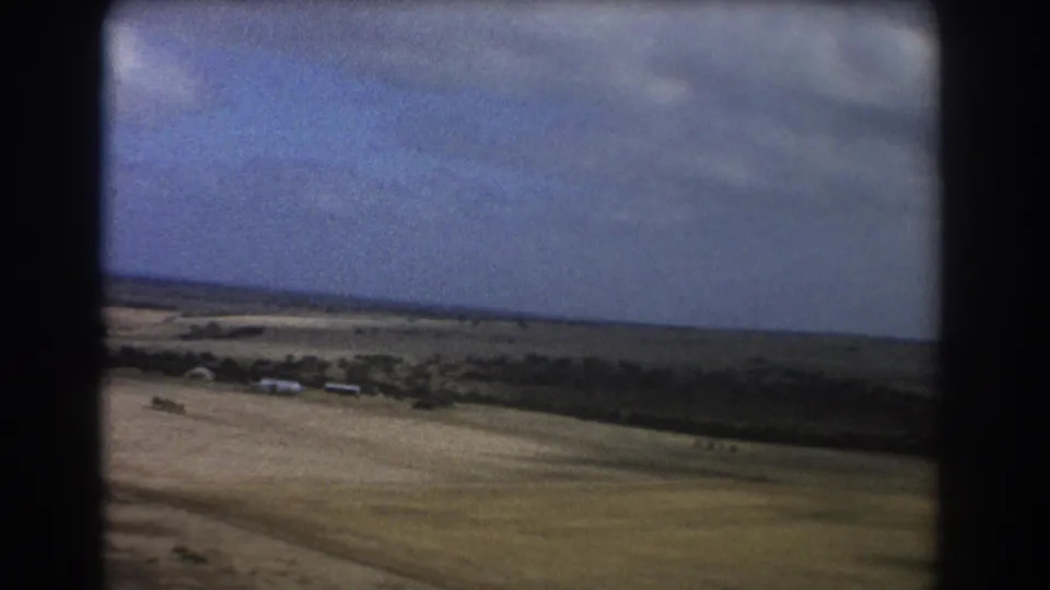 1967 Perth-area home-movie frame of open sandy terrain, scattered greenery, a dark tree line, and small structures near the horizon.