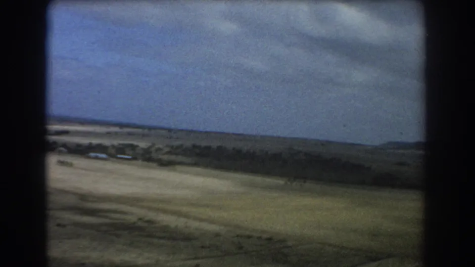 Aerial 8mm frame near Perth with pale ground, scrub, distant low hills, and a wide cloudy sky.