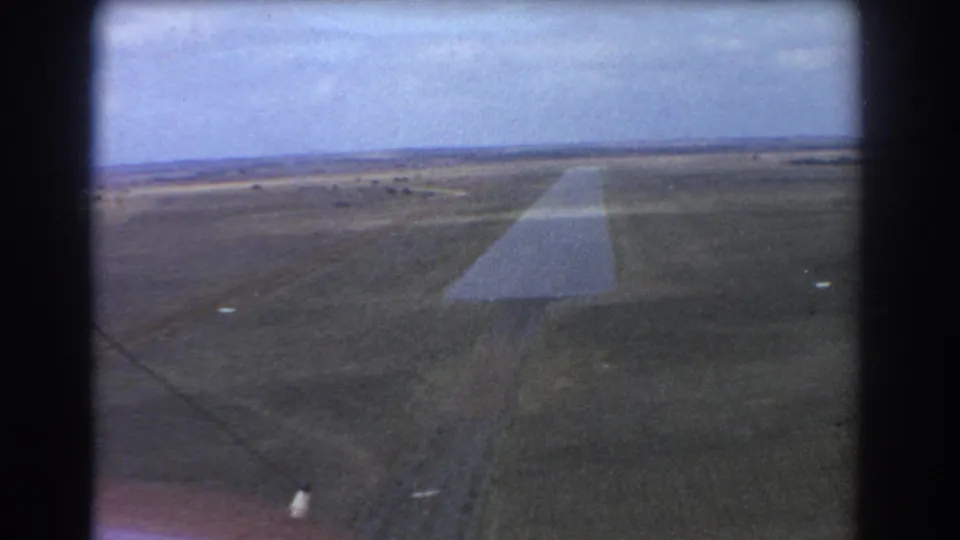 Aerial home-movie frame near Perth showing a runway, surrounding fields, distant horizon, and soft 8mm texture.