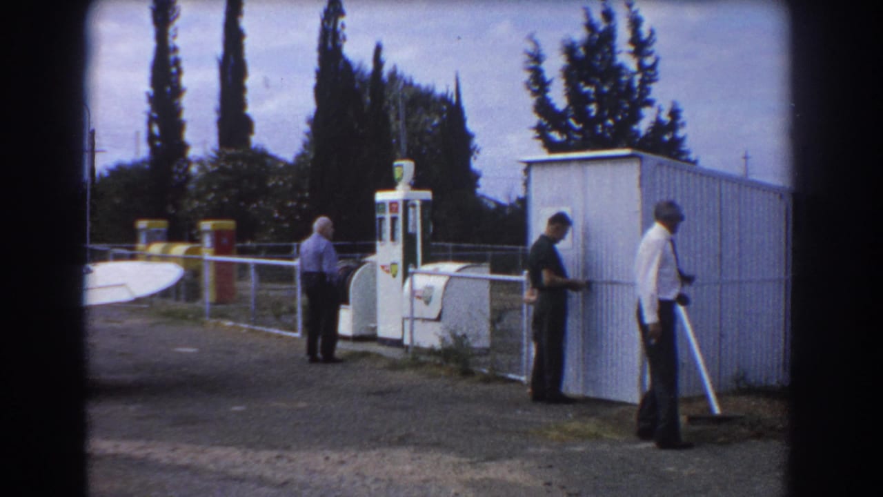 At a Perth airfield in 1967, three adult men stand near a white fuel pump, corrugated shed, railing, and partial aircraft tail.
