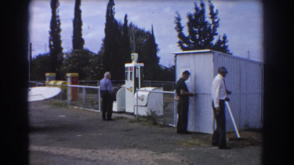 At a Perth airfield in 1967, three adult men stand near a white fuel pump, corrugated shed, railing, and partial aircraft tail.