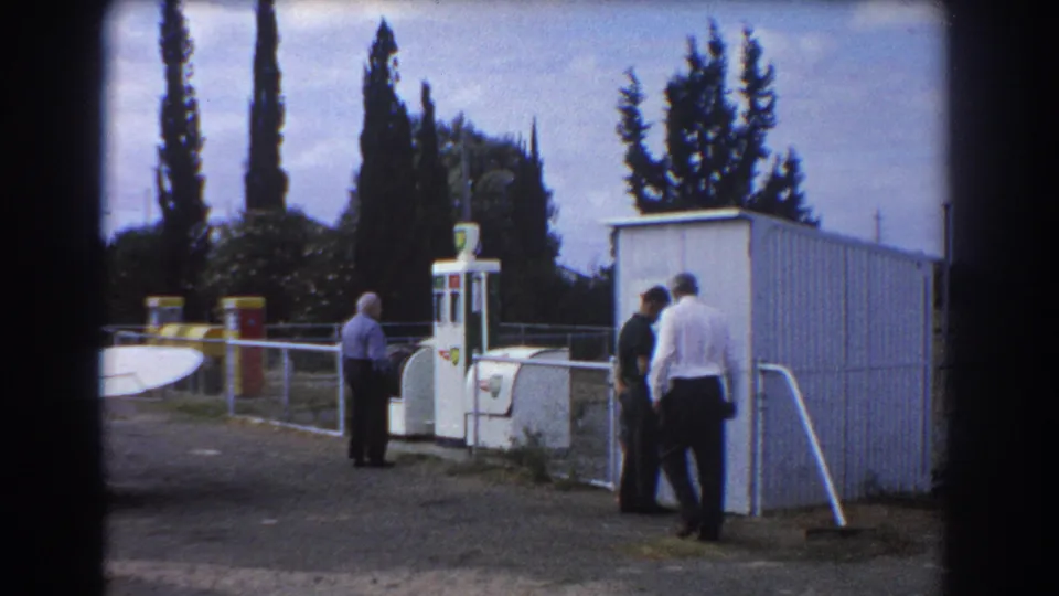 Three adult men appear by a white airfield pump and corrugated shed in this 1967 Perth home-movie frame.