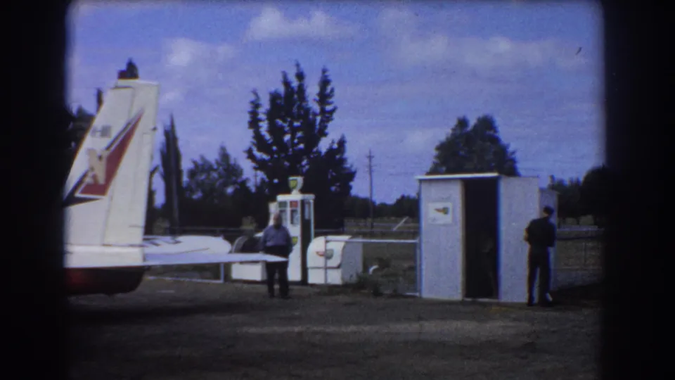 A white aircraft tail with a colored marking enters the Perth airfield frame as men stand near the pump and shed.
