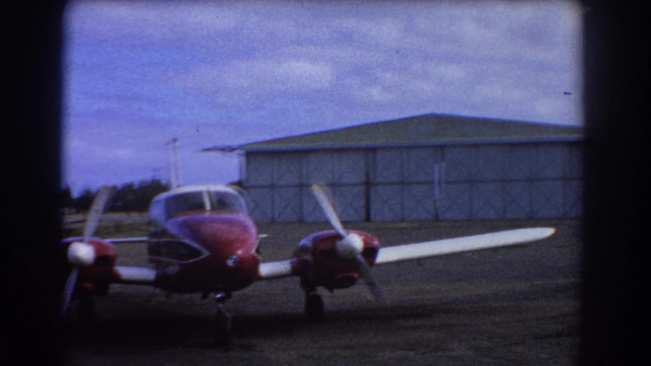 Red and white light aircraft parked before a metal hangar under cloudy sky.