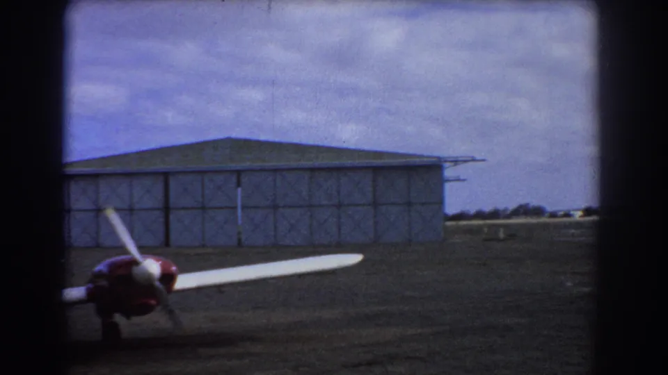 1967 Perth frame with part of a red and white light aircraft in the foreground and a metal hangar across the airfield.