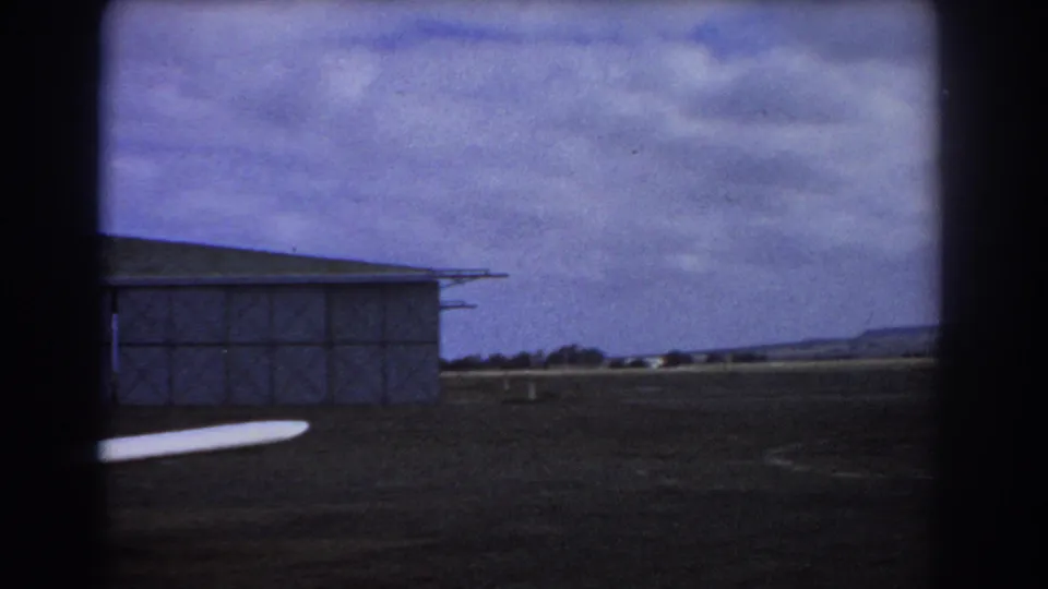 1967 Perth home-movie frame showing the aircraft wing at lower left, a partial hangar, open ground, and distant low hills.