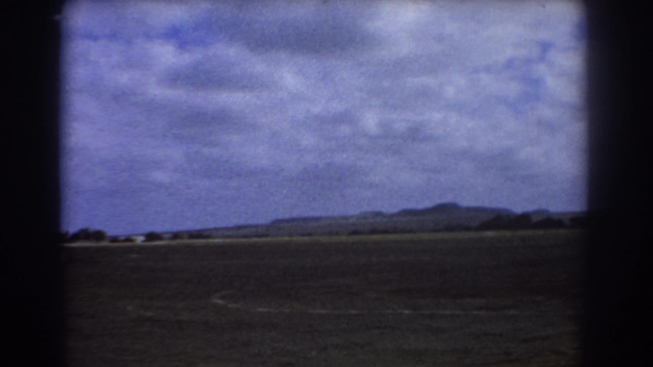 Flat landscape with distant hills under a cloudy sky, framed by dark aircraft-window edges.