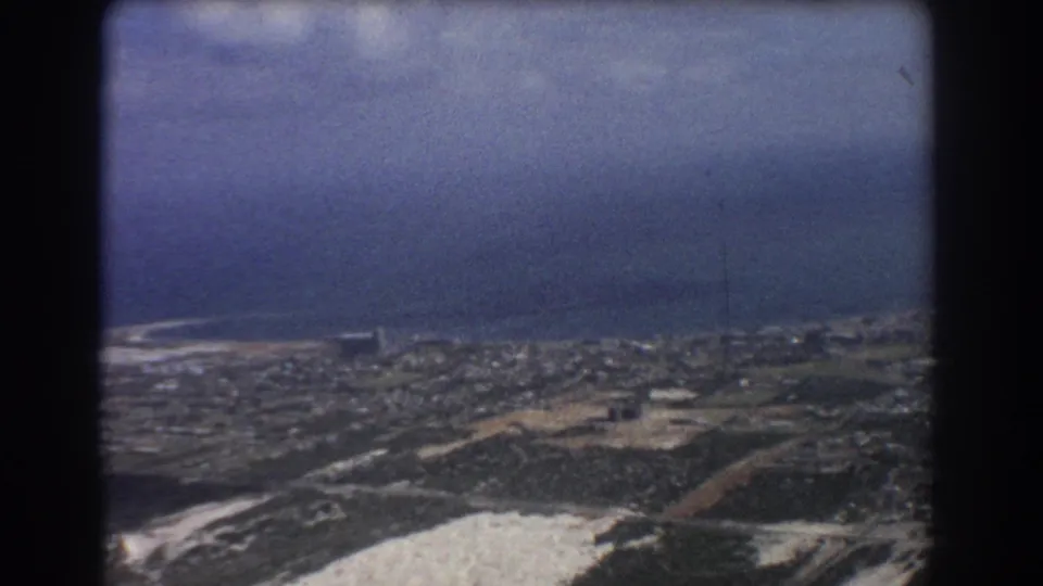 1967 8mm aerial view likely over Perth, Australia, showing urban blocks, open ground, shoreline, and ocean beyond.