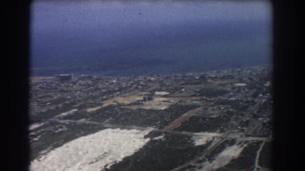 High aerial view over a coastal urban area with roads, fields, and ocean on the horizon.