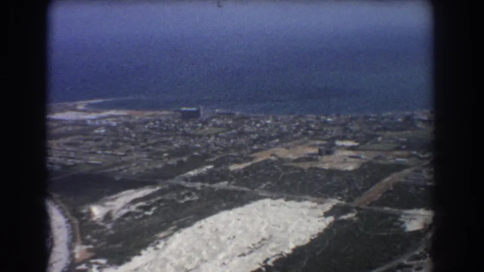 1967 8mm aerial frame likely over Perth, with a sweeping view of shoreline, city blocks, roads, and pale open terrain.