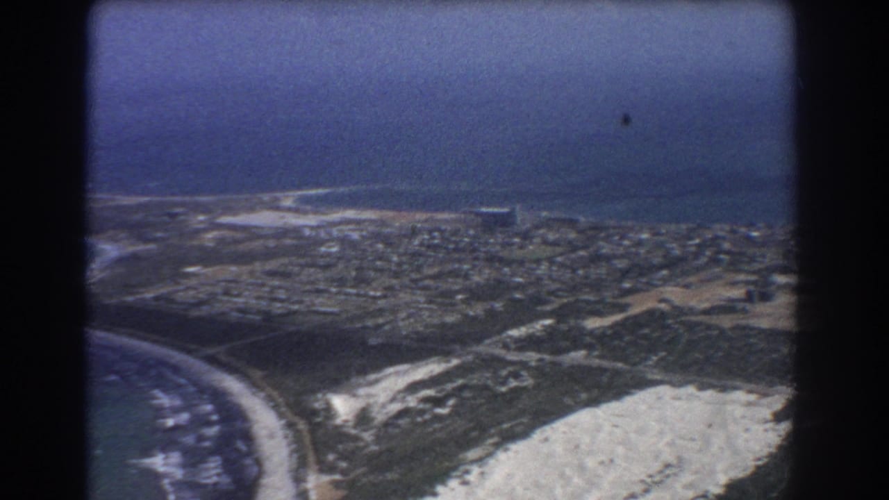 1967 8mm aerial view over Perth coastline with ocean, beach, sandy dunes, roads, and low-rise homes.