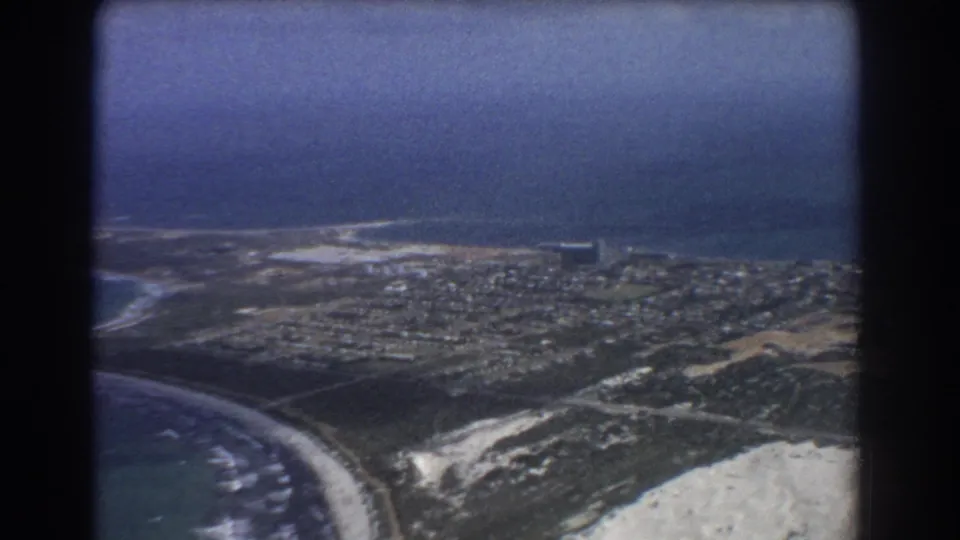 1967 Perth aerial frame showing a curving beach, dune areas, residential streets, and blue ocean beyond.