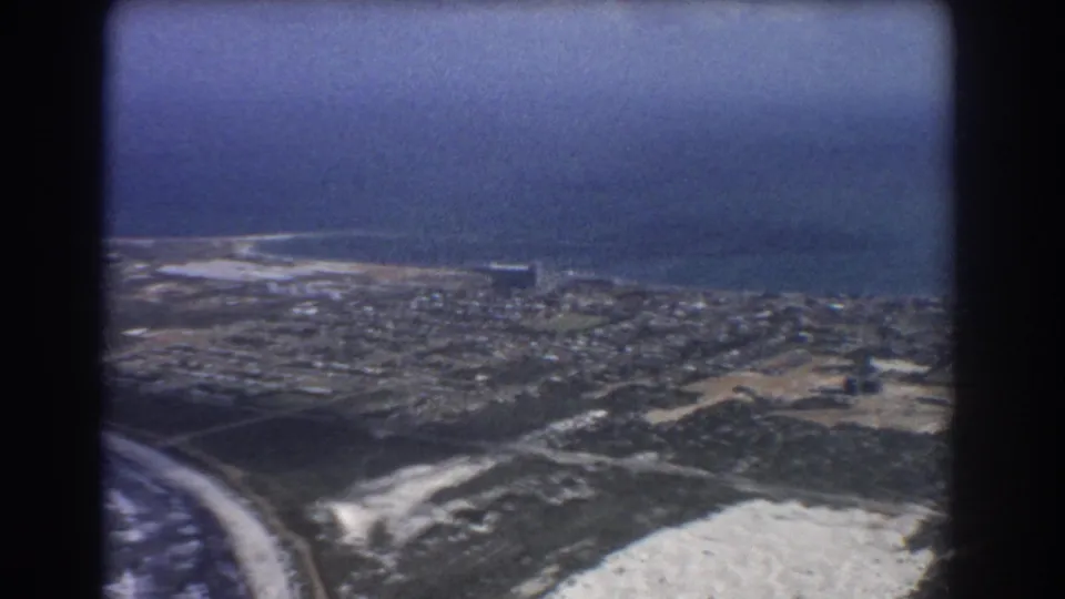 1967 8mm aerial view over Perth's coastal edge, with suburban blocks, sandy ground, beach, and sea under haze.