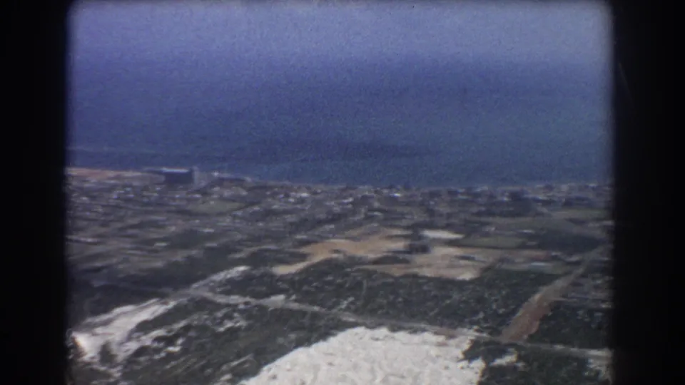 1967 Perth aerial frame with low-rise coastal development, sandy open areas, roads, and ocean stretching across the horizon.