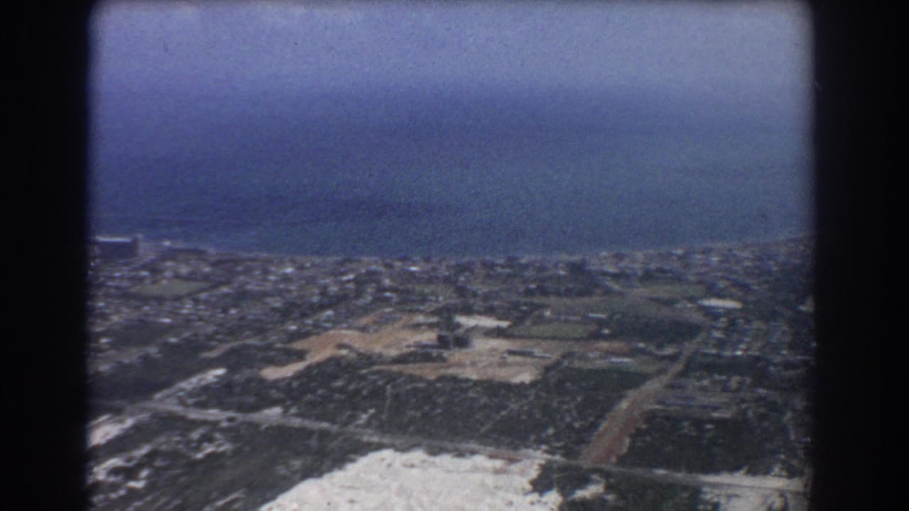 Aerial view of coastline, ocean, suburbs, and open fields seen through dark film-frame edges.