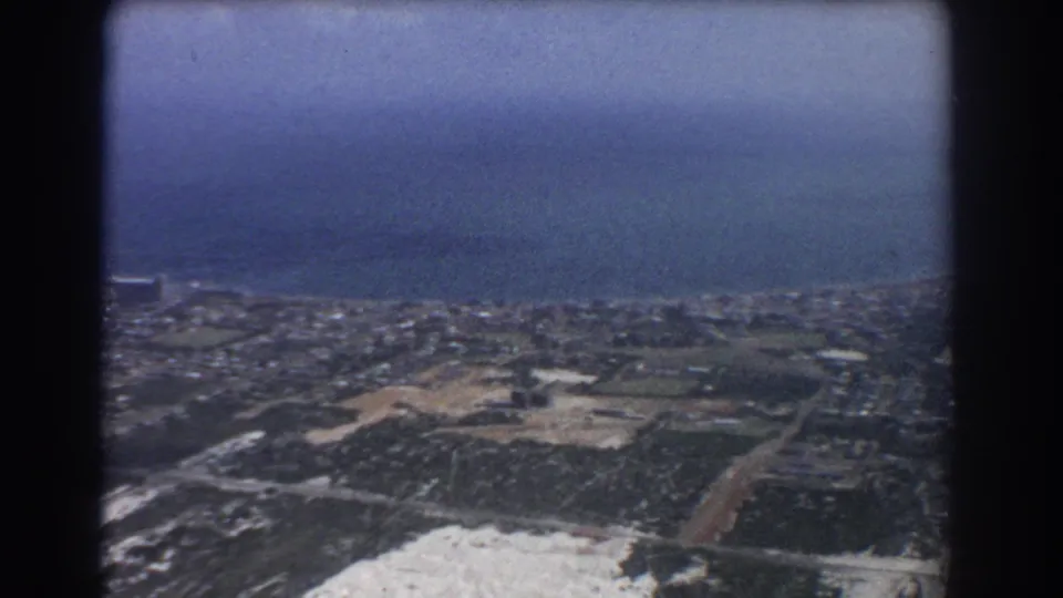 1967 8mm aerial view over Perth, Australia, showing ocean, suburban streets, fields, sandy ground, and a hazy coastal horizon.