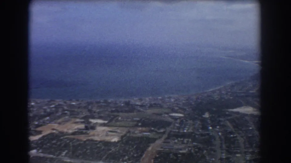 1967 aerial footage over Perth shows a wide ocean horizon, coastal land, suburban neighborhoods, fields, and roads from above.