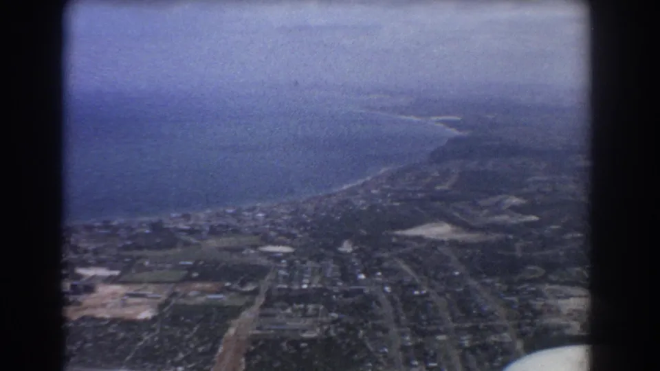 Perth, Australia, in 1967 from above, with ocean along the coast, suburban streets, green fields, and undeveloped patches visible through soft 8mm grain.