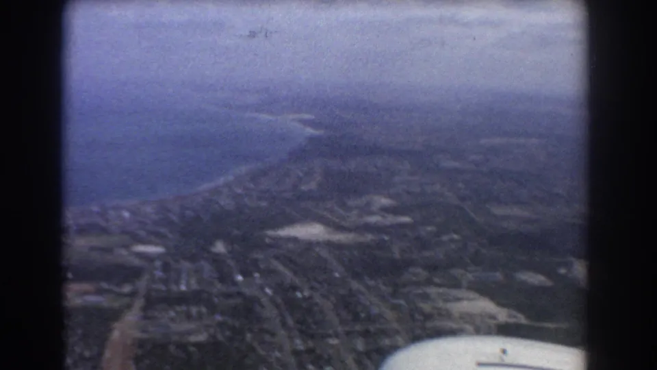 Late-1960s 8mm aerial view over Perth coastline, with ocean, suburbs, fields, haze, and what appears to be part of an aircraft edge at lower right.