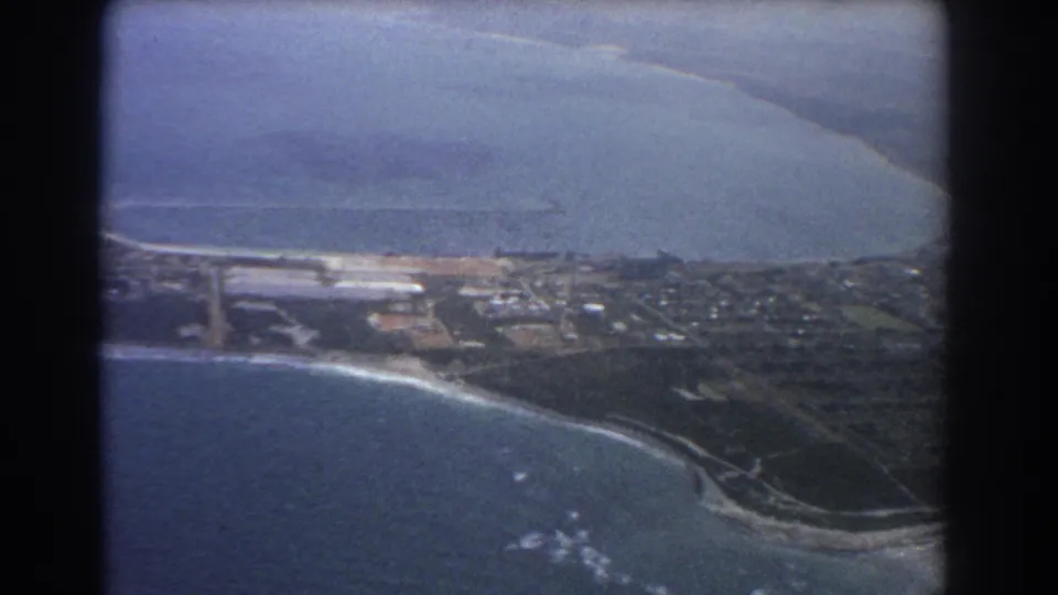 1967 Perth aerial frame with coastline, pale beach, coastal development, roads, ocean water, and inland landforms visible below.