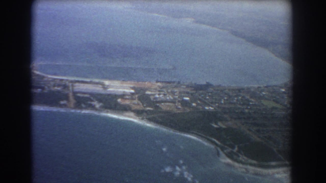 Aerial view of ocean, curved shoreline, roads, buildings, and green coastal land.