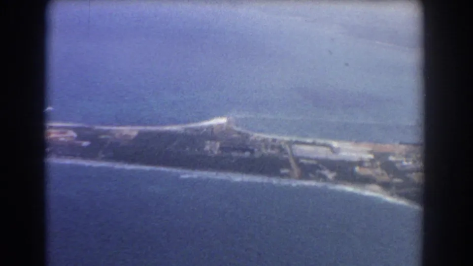 1967 Perth aerial frame with ocean foreground, pale shoreline, dark coastal land, and low structures in a faded 8mm image.