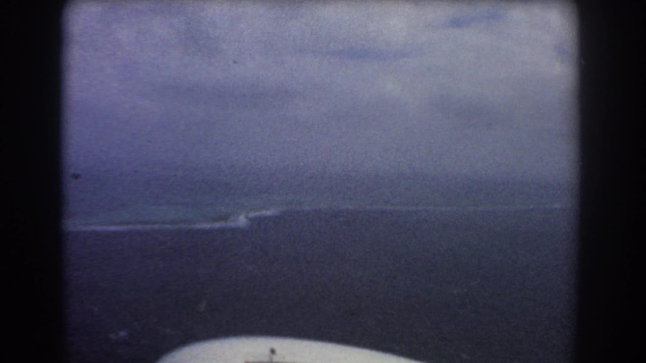 Aerial view of dark ocean, cloudy sky, and a pale shoreline seen through an aircraft window.