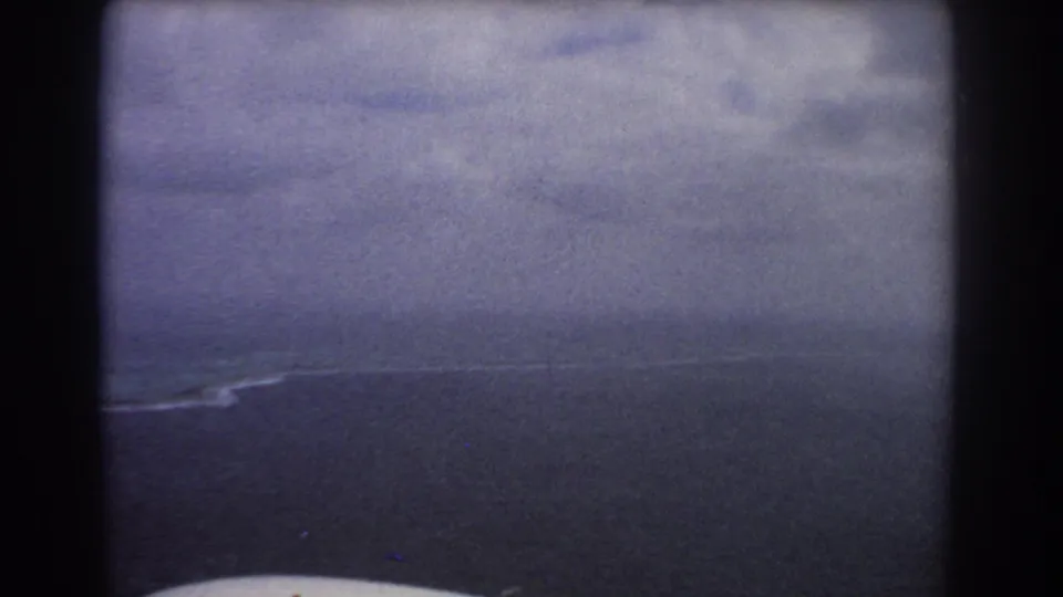 Aerial 8mm frame over the Perth coastline, showing dark water, overcast sky, a thin surf line, and aircraft-window borders.