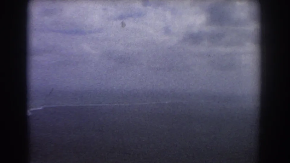 1967 home-movie aerial frame with cloud cover, dark sea, a small strip of surf, and heavy aircraft-window framing.