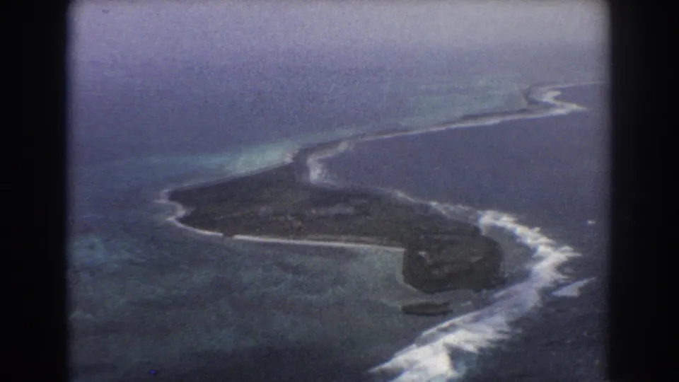 The aerial view continues over the island and reef, with dark land, pale surf, and blue ocean in a soft 8mm frame.