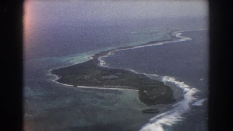 Faded 8mm aerial frame shows the island shoreline, white wave breaks, and shallow turquoise water around the reef.