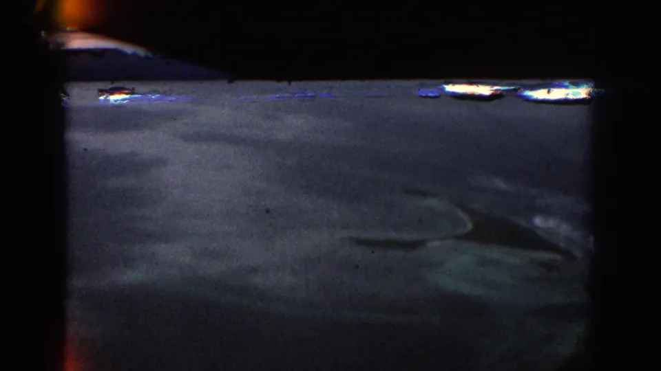 A darker 8mm frame partially shows ocean shallows, reef markings, and the edge of the island beneath film-frame shadow.