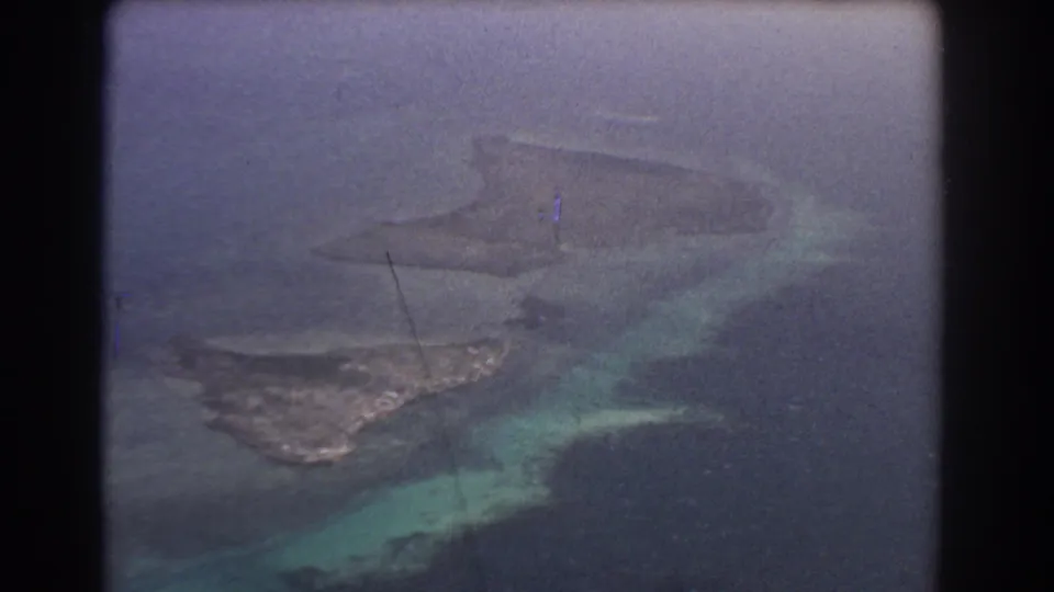 1967 8mm aerial view showing low coastal islands or sandbars in blue-green water, with soft focus and aircraft-window framing.