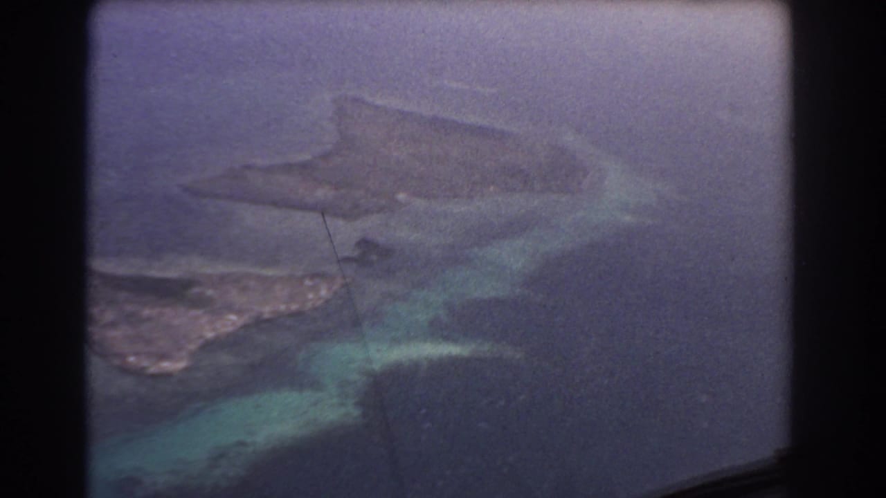 Aerial view through an airplane window of reef, shallow turquoise water, and an island coastline.