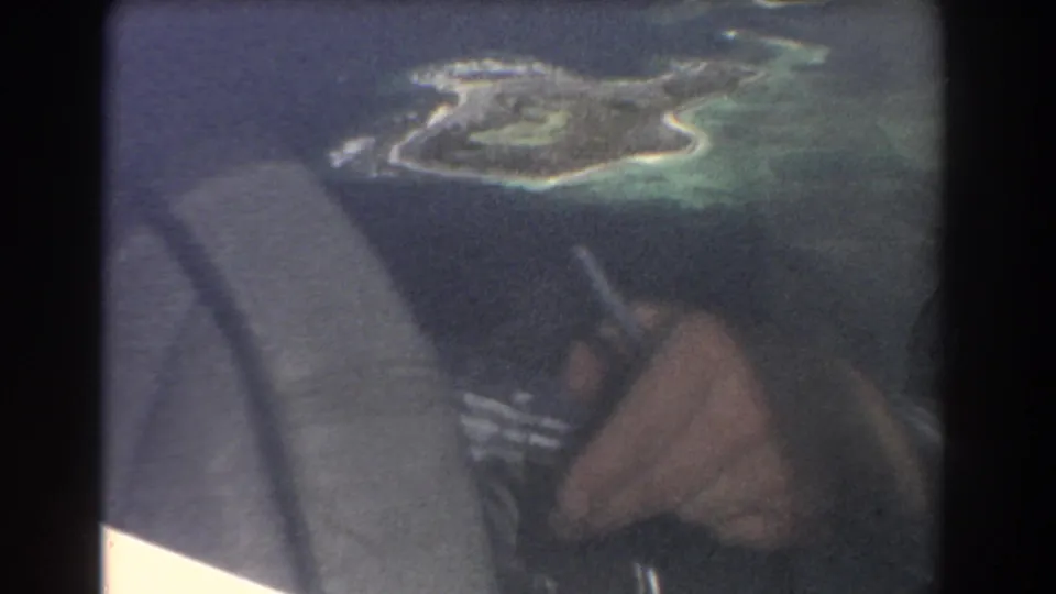 Aerial island and turquoise lagoon visible from an airplane cabin, with a passenger hand and seatbelt in the foreground.
